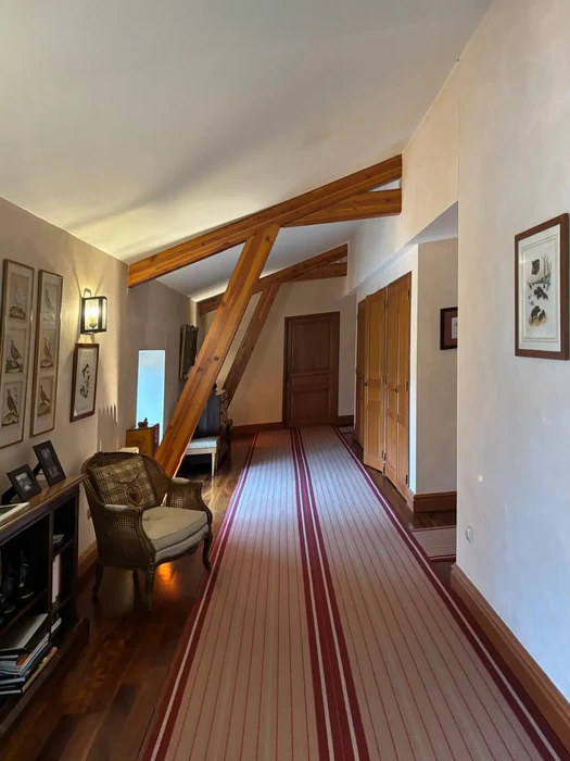 Intérieur résidentiel avec couloir principal présentant un parquet à bandes rouges contrastées, poutres apparentes en bois massif, murs blancs ornés de cadres botaniques, fauteuil en rotin et éclairage d'ambiance tamisé