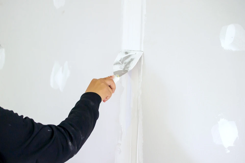 Une femme en tenue de travail rouge et casque de sécurité blanc applique un enduit sur un mur intérieur avec une taloche